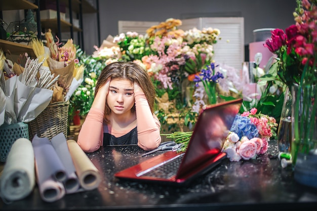 걱정 된 젊은 꽃집 머리에 손을 잡고 노트북을 봐. 테이블에 종이 롤이 다시 있습니다. 그녀의 뒤에는 화려한 꽃과 식물이 많이 있습니다.