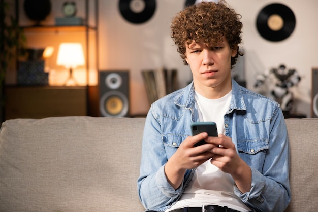 A worried young man in a denim shirt sits on the couch in the living room and looks at the camera