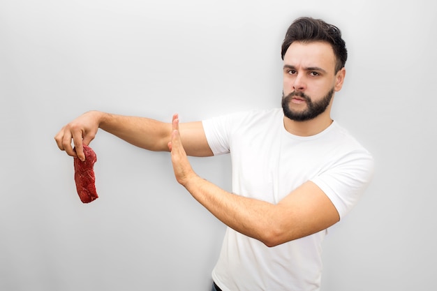 Worried young man stands and holds piece of meat. It is bad. Young man shows stop symbol with hand. He looks and poses.