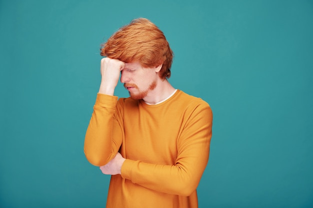 Worried young redhead man with beard focused on mind rubbing forehead with fist on blue