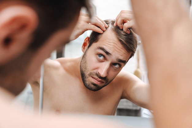 Worried young shirtless man examining his hair while looking at the bathroom mirror