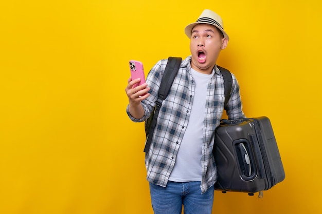 Worried young traveler tourist Asian man 20 years old wears casual clothes hat with backpack holding suitcase and mobile phone looking aside isolated on yellow background Air flight journey concept