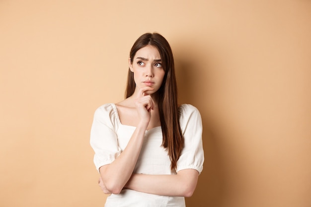 worried young woman staring pensive aside, touching chin and thinking, pondering plan, looking for ideas, standing on beige background.