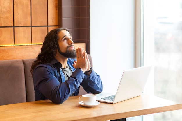 Worry handsome young adult man freelancer in casual style sitting in cafe and talking with his friend in laptopholding hand like pray and looking up Indoor lifestyle concept