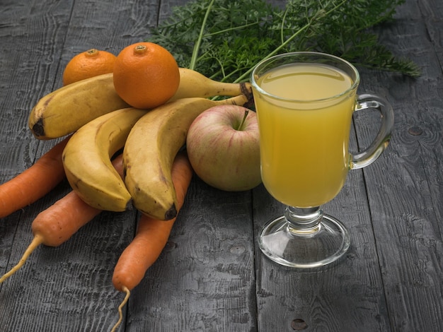 Wortelen, bananen, appels, mandarijnen en een glas smoothies op een houten tafel