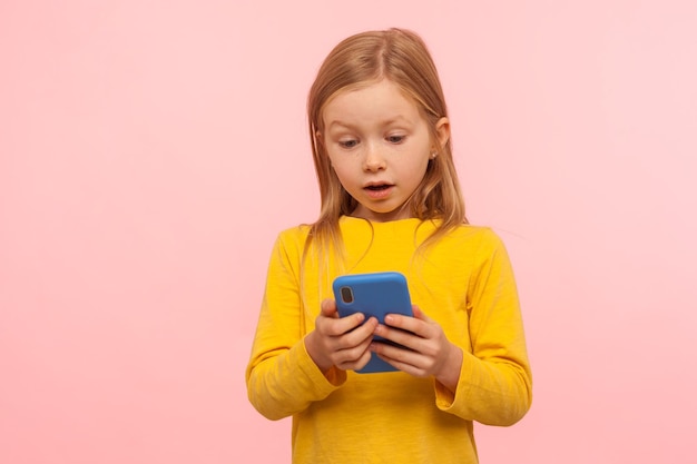 Wow amazing app for kids Portrait of astonished little girl using smartphone with shocked expression playing game on mobile device pleasantly surprised by easy application studio shot isolated