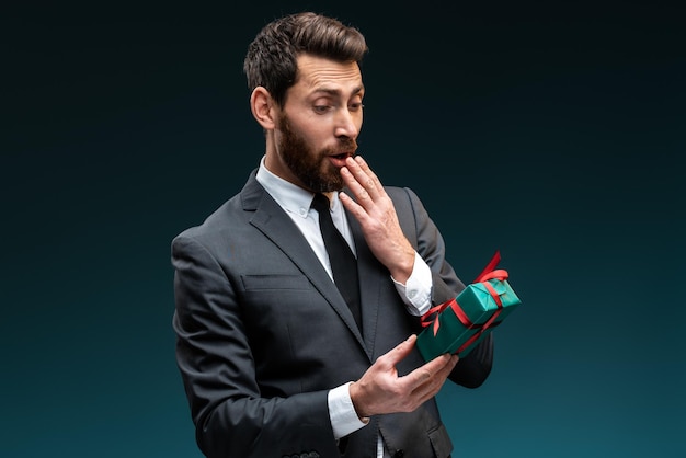 Wow, amazing present. Portrait of shocked man in formal suit looking at the gift box with open mouth and feeling surprised. Indoor studio shot isolated on blue background