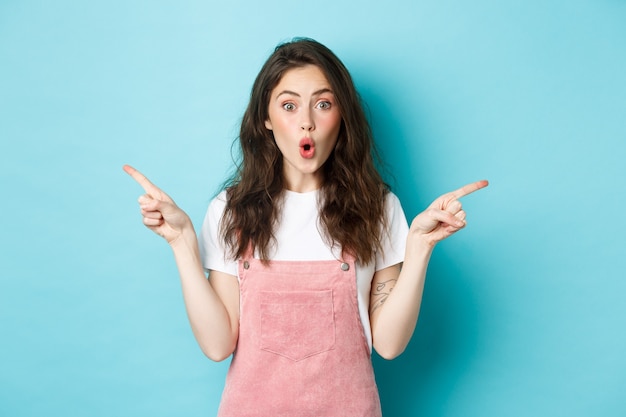 Wow check it out. Excited brunette girl with glamour summer make up, pointing fingers sideways at left and right sides, gasping amazed, standing over blue background.
