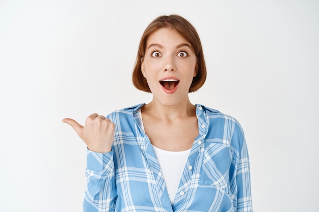 Wow check this out. Amazed and excited young woman open mouth fascinated, pointing left at look astonished , showing way, standing on white wall