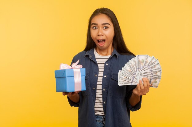 Wow crazy gift shopping portrait of surprised girl in denim shirt holding present box dollar money banknotes and looking amazed shocked by purchase cashback and bank loan studio shot isolated