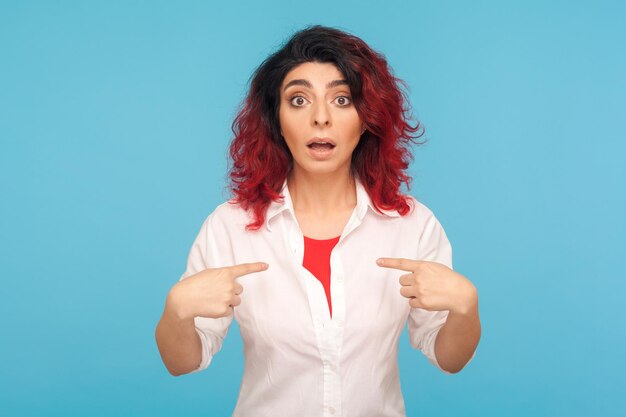 Wow, dit ben ik Portret van verbaasde hipster vrouw met rood haar staren in verbazing naar de camera en wijzend zichzelf geschokt door persoonlijk succes indoor studio shot geïsoleerd op blauwe achtergrond