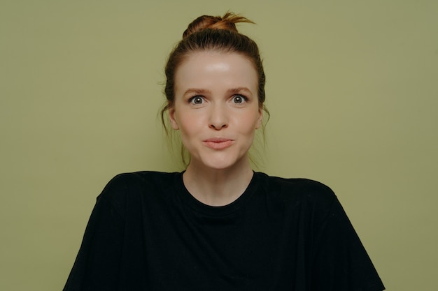 Wow emotion. Wide eyed young woman in black tshirt with hair in bun expressing surprise over what just happened, staring straight at camera, isolated in front of green background