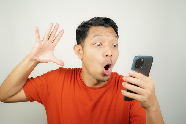 Wow face of Asian man in orange tshirt shocked what he see in the smartphone on isolated background