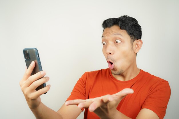 Wow face of Asian man in orange tshirt shocked what he see in the smartphone on isolated background