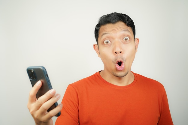 Photo wow face of asian man in orange tshirt shocked what he see in the smartphone on isolated background