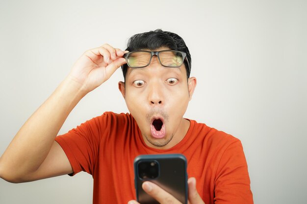 wow face of asian man in orange tshirt shocked what he see in the smartphone on isolated background
