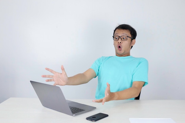 젊은 아시아 남자의 와우 얼굴은 파란색 셔츠를 입고 고립된 회색 배경에서 작업할 때 노트북에서 보는 것에 충격을 받았습니다.