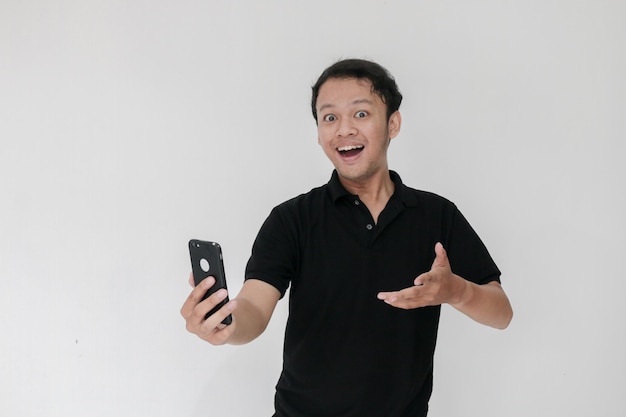 Вау, лицо вашего азиатского мужчины шокировано тем, что он видит в смартфоне на изолированном сером фоне Индонезия Мужчина носит черную рубашку Изолированный серый фон