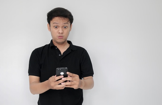 Wow faccia del tuo uomo asiatico ha scioccato ciò che vede nello smartphone su sfondo grigio isolato indonesia uomo indossa una camicia nera sfondo grigio isolato