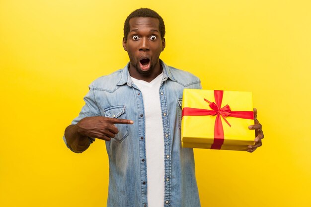 Wow geweldig cadeau portret van geschokte man in denim casual shirt camera kijken met open mond en wijzend op geschenkdoos verrast met bonussen indoor studio shot geïsoleerd op gele achtergrond