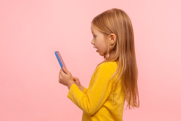 Wow geweldige app voor kinderen Zijaanzicht van verbaasd meisje met smartphone met geschokte uitdrukking speelspel op mobiel apparaat aangenaam verrast door eenvoudige applicatie studio-opname geïsoleerd