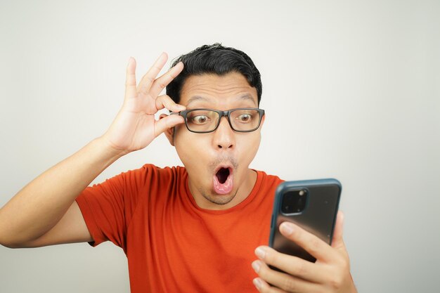 Wow gezicht van Aziatische man in oranje T-shirt geschokt wat hij ziet in de smartphone op geïsoleerde achtergrond