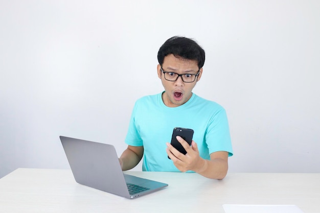 Wow gezicht van jonge aziatische man geschokt door wat hij in de telefoon ziet met laptop ernaast indonesische man met blauw shirt