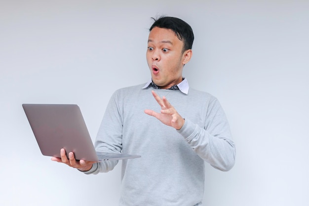 Wow gezicht van jonge Aziatische man geschokt door wat hij op de laptop ziet tijdens het werken op een geïsoleerde grijze achtergrond met een grijs shirt
