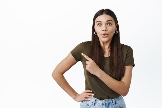 Wow have you seen that Excited young woman pucker lips and stare at camera impressed with store promo deal pointing finger left at copy space white background