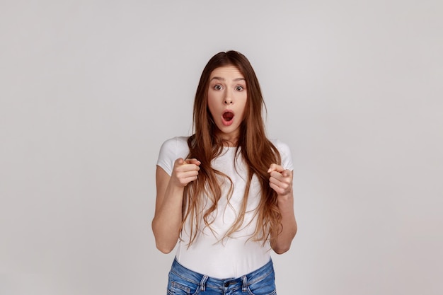 Wow hey you Amazed shocked woman pointing finger to camera looking with surprise noticing and making choice wearing white Tshirt Indoor studio shot isolated on gray background