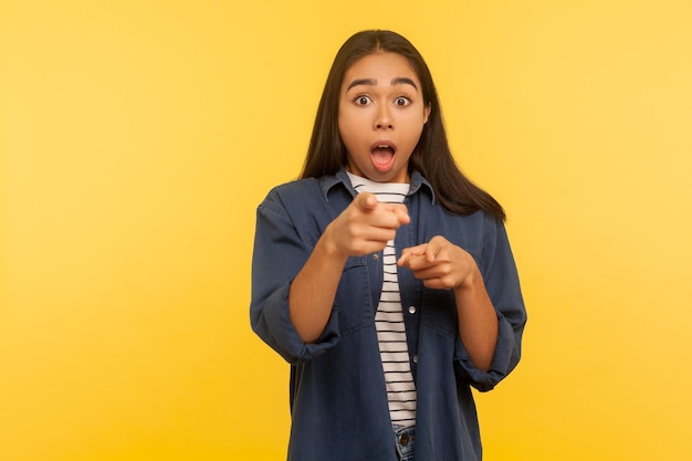 Wow hey you Portrait of surprised shocked girl in denim shirt pointing at something crazy unbelievable and looking amazed disbelief reaction indoor studio shot isolated on yellow background