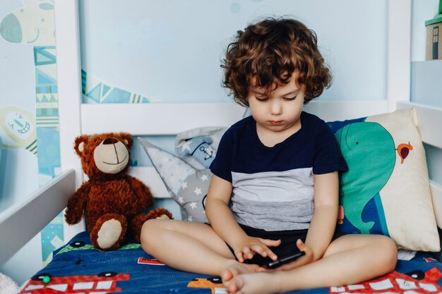 Wow, mi piace quel telefono. bambino con smartphone. ragazzo che si siede nel letto e che gioca con il telefono cellulare. chiamando mia mamma. carino piccolo bambino tiene il cellulare in mano e guardando attentamente lo schermo.