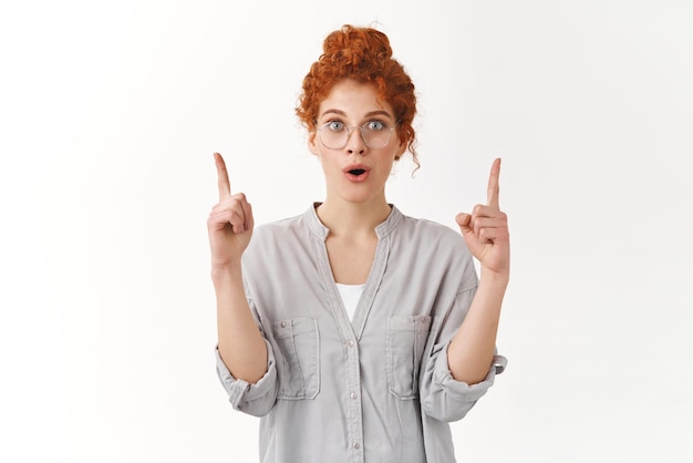 Wow incredible promo checkout Amazed speechless cute redhead woman with curly haircut wear glasses and blouse standing amused open mouth astonished pointing up impressed white background