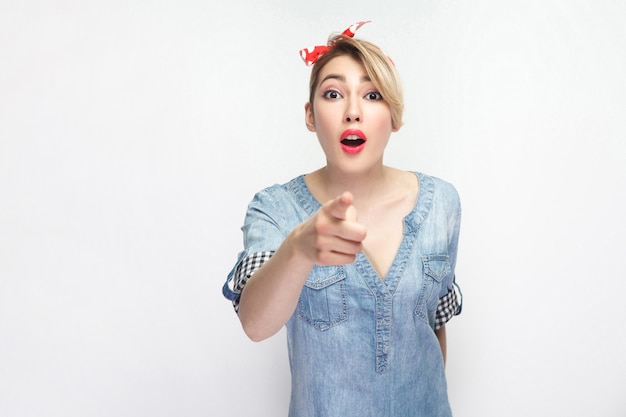 Wow, its unbelievable. Portrait of shocked beautiful young woman in casual blue shirt with makeup and red headband standing and pointing at camera. indoor studio shot, isolated on white background.