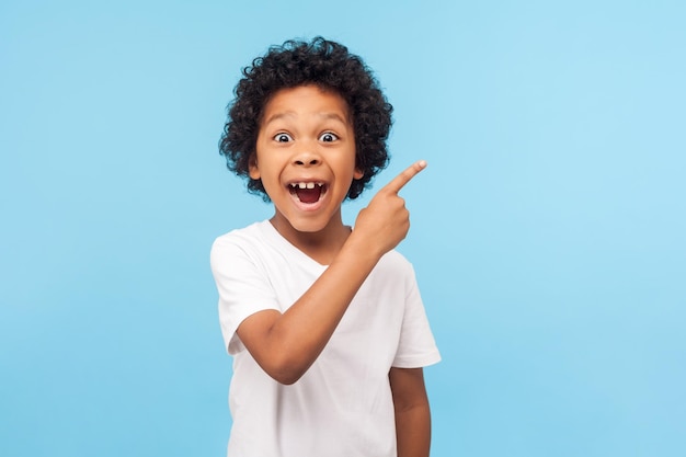 Wow look advertise here Portrait of amazed cute little boy with curly hair pointing to empty place on background surprised preschooler showing copy space for promotional ad indoor studio shot