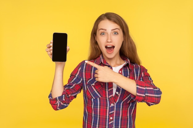 Wow, look at device. Portrait of amazed pretty girl in shirt pointing at cellphone in her hand and expressing shock surprise, advertising of mobile app or gadget. indoor studio shot yellow background