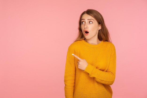 Wow, look. portrait of amazed ginger girl in sweater pointing to the side and looking with surprised face, shocked by your text on copy space, advertising. studio shot isolated on pink background