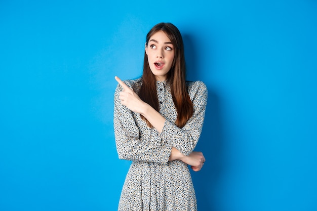 Wow look there. Excited pretty lady in dress looking and pointing left wondered, checking out advertisement, standing on blue.