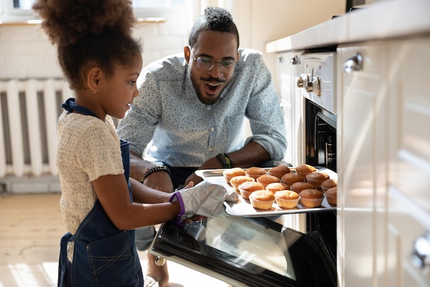 Wow looks great Excited caring millennial african dad or grown elder brother watching concentrated small black daughter or younger preteen sister taking pan with self baked tasty muffins out of oven
