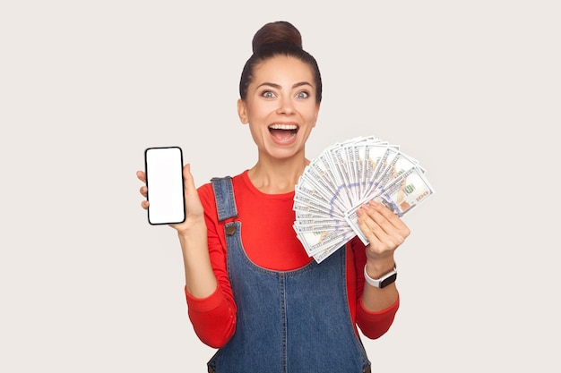 Wow, mobile payment, online shopping. Portrait of amazed stylish pretty girl with hair bun in denim overalls holding cell phone and money, looking shocked. studio shot isolated on white background