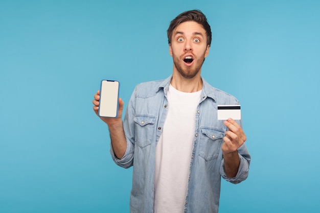 Wow ongelooflijke mobiele winkel-app en online betaling Portret van een verbaasde man in een werknemer denim shirt met mobiele telefoon en creditcard op zoek geschokt studio-opname geïsoleerd op blauwe achtergrond