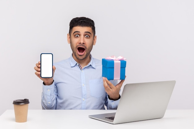 Wow, online shopping advertisement. amazed man office employee sitting at workplace, holding gift box and cell phone with blank mock up display, looking shocked at camera. indoor studio shot isolated