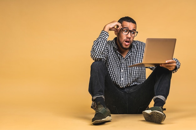 Wow online working concept african american business man\
relaxed working and browsing internet on laptop computer freelancer\
sitting on floor and typing isolated over beige