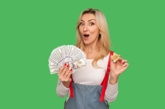 Wow profitable digital investment Portrait of amazed adult woman in stylish overalls holding dollars and btc coin showing bitcoin money vs cash indoor studio shot isolated on green background