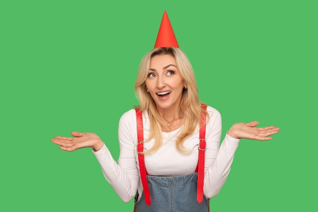 Wow, surprise. Portrait of amazed beautiful woman with funny cone on head standing with raised hands and open mouth, shocked by unexpected birthday party. studio shot isolated on green background