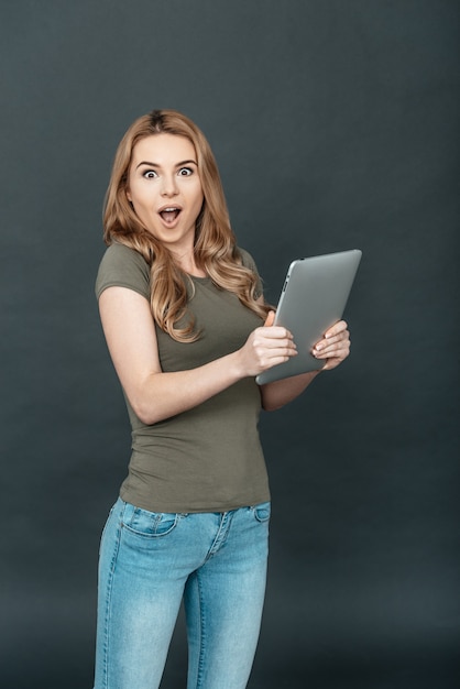 Wow! Surprised young woman with blond hair holding digital tablet with opened mouth 