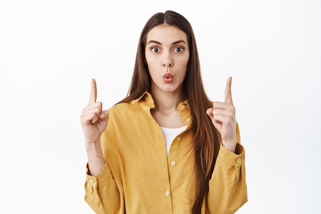 Wow thats interesting, look above. Curious young woman pucker lips and raise eyebrows surprised, looking intrigued, pointing fingers up at copy space with logo, standing over white background