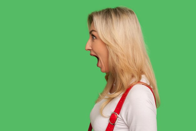 Photo wow unbelievable side view of astonished adult blond woman standing with wide open mouth and shouting in surprise shocked expression indoor studio shot isolated on green background copy space