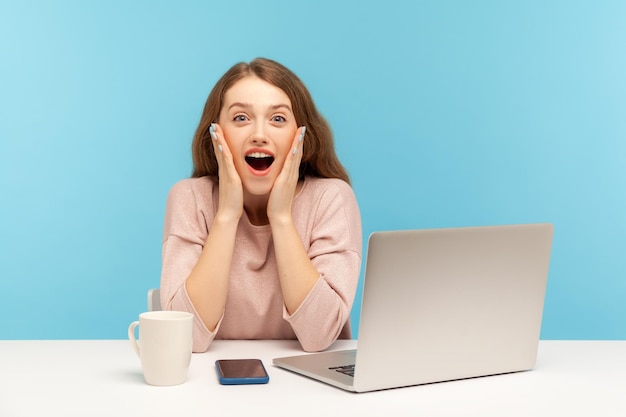 Wow unbelievable Surprised astonished young woman employee sitting at workplace with open mouth shouting in amazement shocked by unexpected news indoor studio shot isolated on blue background