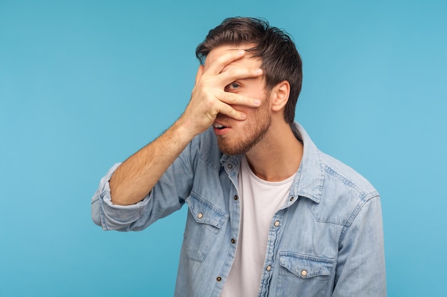 Wow, cos'è? ritratto di curioso uomo ficcanaso in camicia di jeans da lavoratore che guarda attraverso le dita con espressione sorpresa e curiosa, guardando contenuto proibito, spionaggio segreto. girato in studio isolato